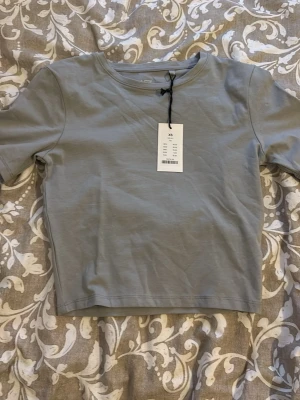 Ljusgrå croppad t-shirt från Arket - Säljer en stilren ljusgrå croppad t-shirt från Lager157. T-shirten har korta ärmar, rund halsringning och är gjord i mjuk bomull. Perfekt basplagg för en clean och enkel look. Storlek Xs. Helt ny med prislapp. 