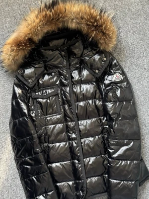 Svart glansig dunjacka Moncler - Snygg svart dunjacka från Moncler med glansigt yttertyg och stor huva med lyxig pälsdetalj. Jackan har bröstficka med knapp, sidofickor och Moncler-logga på ärmen. 