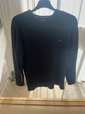 Mörkblå stickad tröja Tommy Hilfiger - Snygg mörkblå stickad tröja från Tommy Hilfiger i storlek M. Tillverkad i en mjuk mix av bomull och silke. Tröjan har rund halsringning och en liten broderad logga på bröstet. Perfekt för en clean och stilren look.