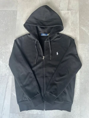 hoodie från Polo Ralph Lauren - Svart hoodie med dragkedja från Polo Ralph Lauren. Klassisk design med huva och dragsko, två fickor framtill och vit broderad logga på bröstet. Tillverkad i mjukt material som är skönt att bära. Perfekt för en avslappnad och stilren look.