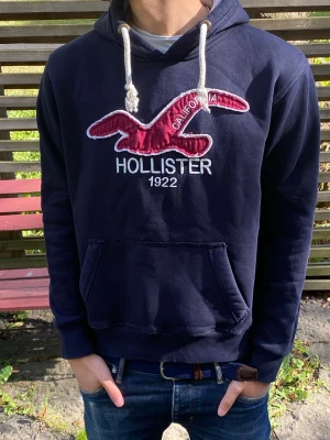 Mörkblå Hollister hoodie med logga - Snygg mörkblå hoodie från Hollister med stor röd broderad logga och texten 'California 1922' på bröstet. Mjuk insida, grått foder i huvan och vita dragsnören. Perfekt för en avslappnad och cool stil.
