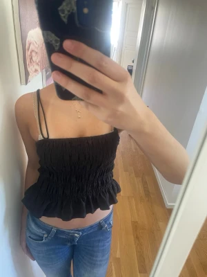 Svart smockad bandeau topp divided H&M  - Svart bandeau topp med smockad design och volangkant nertill. Tunt och lätt material, perfekt för varma dagar. Axelbandslös modell med knytband på sidorna för extra detalj. Snygg och trendig topp som passar till jeans eller kjol.
