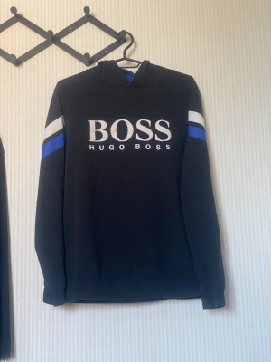 Svart hoodie från Hugo Boss - Svart hoodie från Hugo Boss med stor vit logga på bröstet. Har vita och blå ränder på ärmarna och huva. Perfekt för en stilren och sportig look.