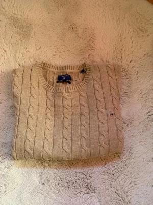 Beige kabelstickad tröja från Gant - Snygg beige kabelstickad tröja från Gant med rund halsringning och diskret logga broderad på bröstet. Tröjan har långa ärmar och ribbade muddar vid ärmslut och nederkant. Perfekt för lager-på-lager och en klassisk look.