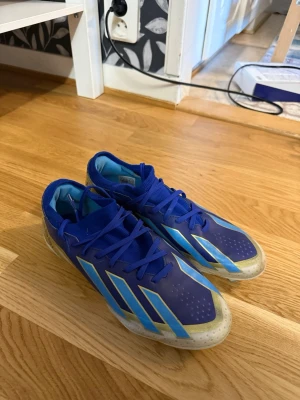 Blå Adidas fotbollsskor Messi - Säljer ett par blå fotbollsskor från Adidas Messi-kollektionen med guld- och ljusblå detaljer. Skorna har snörning, låg profil och dobbar för spel på gräs. Tillverkade i syntetmaterial för bra grepp och kontroll på planen. Storlek 40!