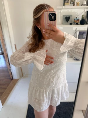 Vit spetsklänning med volang - Superfin vit klänning med spetsdetaljer och volang nertill. Klänningen har långa ärmar med utsvängda ärmslut och ett romantiskt blommönster i tyget. Perfekt för sommarens alla tillfällen! Hade tänkt att ha på studenten men de blev inte av.. så nästintill oanvänd🥰 från Nakd, storlek 40 men mer som 38