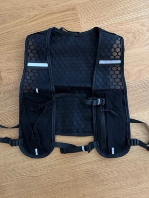 Svart löparväst med reflexdetaljer - Säljer en svart löparväst med meshpaneler och hexagonmönster. Västen har reflexdetaljer framtill, justerbara remmar och flera fickor för småprylar och flaska. Perfekt för löpning eller cykling när du vill ha med dig det viktigaste utan att det är i vägen.