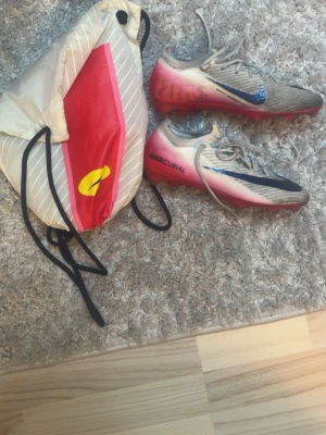 Nike Mercurial elite - Säljer ett par Nike Mercurial fotbollsskor i grått med blå och svarta detaljer samt röda sulor. Skorna har snörning och är tillverkade i syntetmaterial för bästa grepp och känsla på planen. Medföljer en praktisk gympåse med röd och gul detalj.