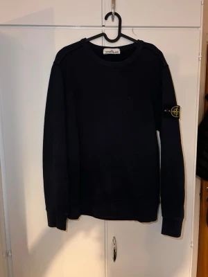 Svart sweatshirt från Stone Island - Snygg svart sweatshirt från Stone Island med klassisk rund hals och ikonisk logotyp-patch på vänster ärm. Tröjan har en enkel och stilren design med ribbade muddar vid ärmslut och nederkant. Perfekt för en avslappnad streetwear-look.