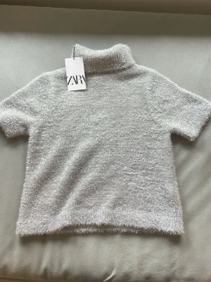 Glittrig silver polotopp från Zara - Supersnygg glittrig polotopp från Zara i silver. Toppen har korta ärmar, mjukt fluffigt material och en hög krage som ger en trendig look. Perfekt för dig som vill sticka ut lite extra och älskar glitter!