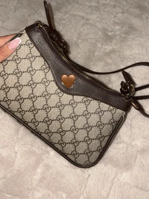  axelväska  - Snygg axelväska i beige och mörkbruna läderdetaljer. Väskan har guldfärgad dragkedja och justerbar axelrem. Perfekt storlek för det viktigaste och en ikonisk accessoar för din outfit. Kedjan kan ibland inte stängas helt om man drar för hårt men annars inget konstigt 