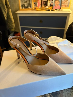 Beige pumps från By Malene Birger - Snygga oanvända beige pumps från By Malene Birger, köpta för ca 5000kr med spetsig tå och smal klack. Skorna har en elegant slingback-design med justerbart spänne och är tillverkade i mjukt mockamaterial. Perfekt för dig som vill ha en stilren och trendig look.