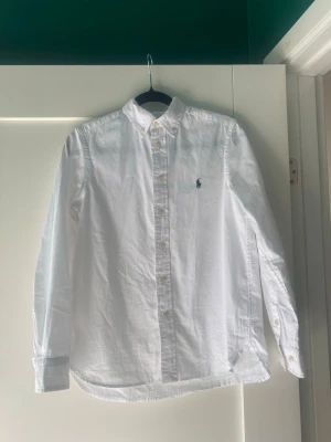 Vit skjorta från Polo Ralph Lauren - Klassisk vit skjorta från Polo Ralph Lauren i slim fit-modell. Skjortan har långa ärmar, knappar framtill och den ikoniska broderade loggan på bröstet. Perfekt för en clean och stilren look.  För barn: storlek 14 år, längd 160. 