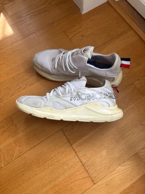 Vita Moncler sneakers med logga - Snygga vita sneakers från Moncler med tydlig logga på sidan och klassisk röd, vit och blå detalj på hälen. Skorna har en chunky sula, mesh och mockadetaljer samt vita skosnören. Perfekta för dig som gillar streetstyle och exklusiva märken.