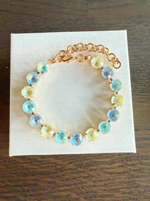 Guldarmband blå/gul  - Armband i guldton med runda stenar  i pastellblått, ljusgult. Pärlorna har en skimrande yta och sitter i en kedja med justerbar längd och karbinlås. Perfekt för dig som gillar färg och vill ha något som sticker ut. Bild 2 är en inspirationsbild hur du kan kombinera armbandet med egna smycken, tex från Svedbom. 