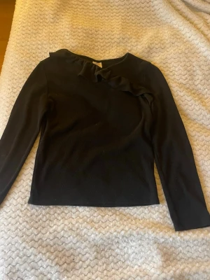 Svart topp med volangdetalj - Svart långärmad topp med snygg volang längs halsringningen. Enkel och stilren design som passar till många outfits. Mjuk och bekväm, perfekt för dig som gillar en klassisk look med en twist.