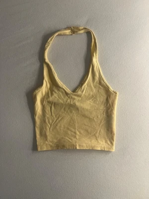 Gul croppad linnetopp med v-ringning - Trendig gul croppad linnetopp med v-ringning och halterneck. Toppen är ärmlös och tillverkad i mjuk bomull, perfekt för varma dagar. Enkel och stilren design som är lätt att matcha med jeans eller kjol.