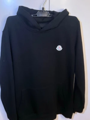 Svart hoodie från Moncler - Svart hoodie/ Moncler med klassisk huva och känguruficka framtill. Diskret Moncler-logga på bröstet. Mjuk och skön bomullsblandning, perfekt för en avslappnad och stilren look.
