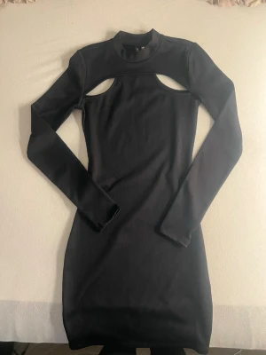 Svart ribbad klänning med cut out - Trendig svart ribbad klänning med lång ärm och cut out-detalj över bröstet. Klänningen har en kroppsnära passform och hög hals, vilket ger en cool och modern look. Perfekt för dig som vill sticka ut lite extra.