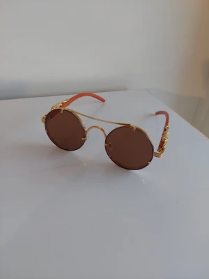 Runda solglasögon med gulddetaljer - Unika solglasögon med runda bruna glas och guldfärgad metallbåge. Skalmarna är orange med snygga gulddetaljer som ger en cool och trendig look. Perfekta för dig som vill sticka ut med accessoarer som har retrovibbar.