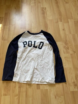 Vit och marinblå långärmad POLO  - Snygg långärmad topp från Polo med vit kropp och marinblå ärmar. Klassisk baseball-inspirerad design med stor POLO-text framtill. Tillverkad i mjuk bomull, perfekt för en avslappnad och sportig stil. Storlek M (10-12)