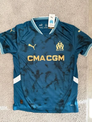 Marseille fotbollströja Puma blå - Snygg blå fotbollströja från Puma för Olympique de Marseille med guldfärgade loggor och tryck. Tröjan har korta ärmar, V-ringning och svarta marmorliknande detaljer. Materialet är lätt och andas, perfekt för träning eller match.