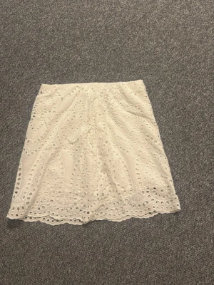 Vit kortkjol med broderade spetsdetaljer - Supersöt vit kortkjol med broderade spetsdetaljer och vågig kant. Kjolen har ett fint hålmönster och elastisk midja för skön passform. Perfekt för dig som gillar romantisk och somrig stil.