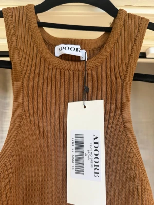 Ribbad långklänning från Adoore - Snygg ribbad långklänning från Adoore i en varm brun färg. Klänningen är ärmlös med rund halsringning och har en figurnära passform. Perfekt för dig som gillar stilrena och trendiga plagg.