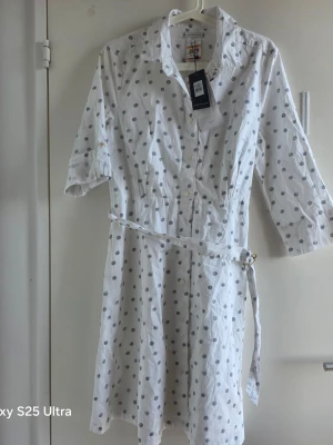Vit prickig skjortklänning Tommy Hilfiger - Vit skjortklänning från Tommy Hilfiger med svarta prickar, trekvartsärm och knappar framtill. Klänningen har ett bälte i midjan och klassisk krage. Perfekt för en fräsch och avslappnad look.