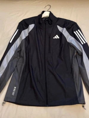 Svart vindjacka från Adidas - Snygg svart vindjacka från Adidas med grå detaljer och vita ränder på ärmarna. Jackan har hel dragkedja, hög krage och en bröstficka med dragkedja. Materialet är lätt och vindtätt, perfekt för aktiva dagar utomhus. Adidas-logga på bröstet och texten 'WIND.DRY' på nederkanten.