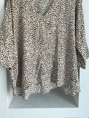 Mönstrad blus från Amelie & Me - Säljer en luftig blus från Amelie & Me i storlek L. Blusen har ett abstrakt mönster i beige, rosa, svart och olivgrönt, v-ringning med dragkedja och vida, korta ärmar. Perfekt för dig som gillar en avslappnad men trendig stil.