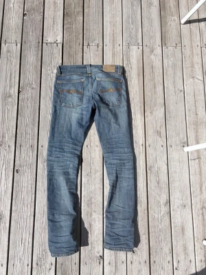 Blå slim fit jeans från Nudie Jeans - Snygga blå slim fit jeans från Nudie Jeans med klassisk femficksdesign och slitningar på ena benet. Jeansen har en ljus tvätt och detaljerade sömmar på bakfickorna. Perfekta för en avslappnad och trendig stil. W28 L32