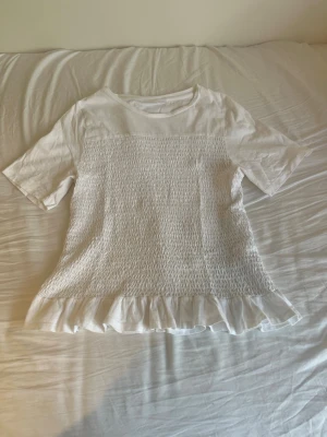 Vit topp med volangkant - Söt vit topp med korta ärmar och smockad front. Toppen har en volangkant längst ner som ger en extra feminin touch. Perfekt till jeans eller kjol för en fräsch look. Materialet känns mjukt och bekvämt. Perfekt nu inför sommaren!☀️
