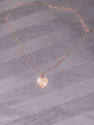 Halsband med hjärthänge i roséguld - Snyggt halsband i roséguldfärgad metall med ett hjärtformat hänge. Hänget har ett stilrent, graverat mönster som ger en cool touch. Perfekt accessoar för att lyfta din outfit med en romantisk vibe.
