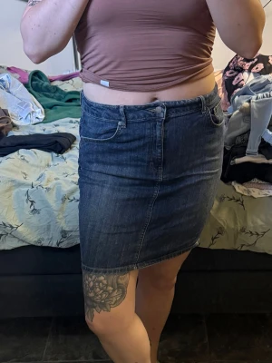 Mörkblå jeanskjol i klassisk modell - Säljer en mörkblå jeanskjol med klassisk femficksdesign och normal passform. Kjolen är kort och har en enkel, tidlös look som passar till det mesta. Perfekt för dig som gillar denim och vill ha en basic kjol i garderoben.