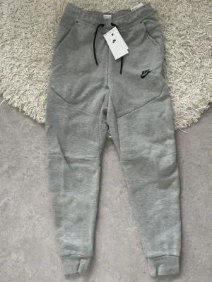 Gråa Nike mjukisbyxor slim fit - Snygga grå mjukisbyxor från Nike med slim fit och tapered ben. Byxorna har svarta dragsnören i midjan, sidofickor och Nike-logga på låret. Perfekta för en avslappnad och sportig stil. Tillverkade i mjukt och bekvämt material.
