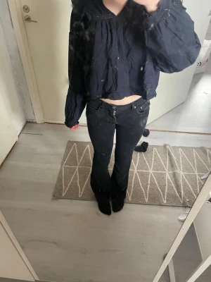 Svart croppad blus med volang - Trendig svart croppad blus med volangkant och rynkade detaljer. Blusen har långa, vida ärmar med resår vid handlederna och dekorativa spetsinslag upptill. Perfekt för dig som gillar en avslappnad men ändå stilsäker look.