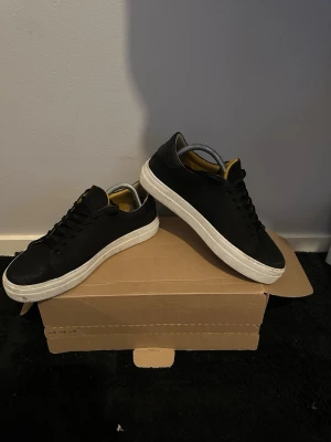 Svarta låga sneakers med vit sula - Snygga svarta sneakers med klassisk vit sula och svarta skosnören. Insidan har en gul detalj och fodret är randigt. Skorna har en stilren och enkel design som passar till det mesta. Materialet ser ut att vara läder eller konstläder.