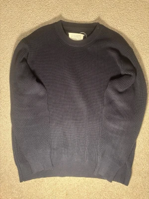 Mörkblå stickad tröja Maison Margiela - Säljer en mörkblå stickad tröja från Maison Margiela. Tröjan har en klassisk rund halsringning, ribbade muddar och är långärmad. Perfekt för dig som gillar minimalistisk och stilren design.