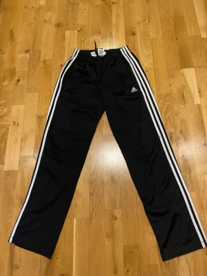 Svarta Adidas mjukisbyxor med ränder stl XSmall bra skick - Svarta mjukisbyxor från Adidas med klassiska vita ränder längs sidorna och Adidas-logga på vänster ben. Byxorna har resår i midjan och raka ben. Perfekta för en sportig och avslappnad stil.