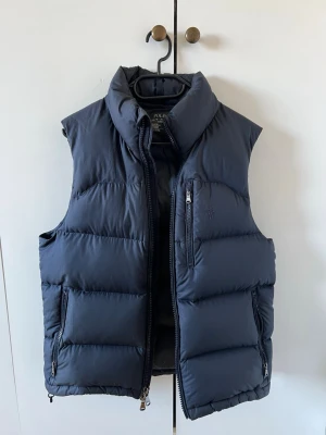 Mörkblå dunväst från Ralph Lauren - Mörkblå dunväst från Polo Ralph Lauren med hög krage och dragkedja framtill. Västen har tre fickor med dragkedja och diskret broderad logga på bröstet. 