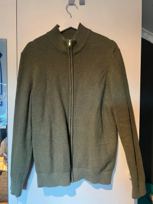 Olivgrön stickad ziptröja East West - Snygg olivgrön stickad ziptröja från East West med hög krage och ribbade muddar. Tröjan har hel dragkedja framtill och är tillverkad i ett grovstickat material som ger en avslappnad men stilren look.