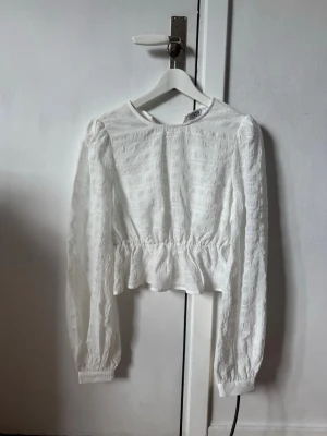Vit blus med volang från NA-KD - Superfin vit blus från NA-KD med långärmad design och volang nertill. Blusen har rund halsringning och är gjord i ett lätt strukturerat tyg som ger en trendig look. Perfekt att styla med jeans eller kjol för en fräsch outfit.