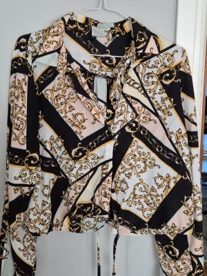 Mönstrad blus från Gina Tricot - Säljer en snygg blus från Gina Tricot med svart, vit och beige botten och guldiga barockinspirerade mönster. Blusen har knytband i halsen, krage och långa ärmar. Materialet känns lätt och flowy, perfekt för att lyfta vilken outfit som helst.