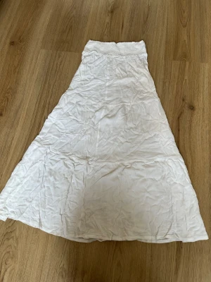 Vit långkjol med smock i midjan - Säljer en vit långkjol med smockad midja och lätt utsvängd passform. Kjolen är enkel och stilren, perfekt för varma dagar. Materialet känns mjukt och luftigt, vilket gör den bekväm att bära.