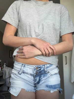 Ljusblå lågmidjade jeansshorts med fransig kant - Säljer ett par ljusblå denimshorts med slitna detaljer och fransig kant. Shortsen har hög midja, tre knappar framtill och klassiska fickor både fram och bak. Perfekta för sommaren och ger en avslappnad vibe.