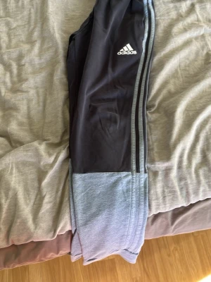 Adidas svarta mjukisbyxor med grå detaljer - Svarta mjukisbyxor från Adidas med grå muddar och klassiska tre ränder längs sidorna. Byxorna har resår i midjan med dragsko och Adidas-logga på låret. Perfekta för chill eller träning, tillverkade i mjukt material.