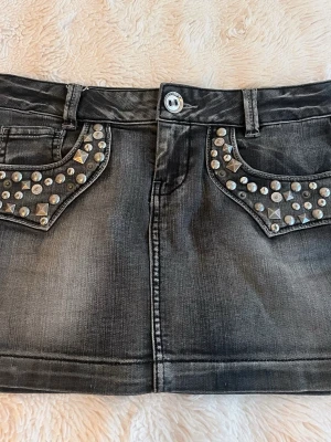 Svart jeanskjol med nitar - Cool svart jeanskjol med slitningseffekt och silverfärgade nitar på både fram- och bakfickor. Kjolen har klassisk femficksdesign, bälteshällor och knappstängning framtill. Perfekt för dig som gillar en edgy stil.