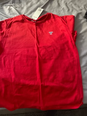 Röd T-shirt från GANT - Röd T-shirt från GANT med klassisk rund hals och liten broderad logga på bröstet. Tillverkad i mjuk bomull som känns skön mot huden. Perfekt basic-plagg med stilren design och snygg passform.