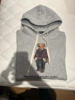 Grå hoodie Polo Bear Ralph Lauren - Snygg grå hoodie från Ralph Lauren med Polo Bear-tryck framtill. Klassisk huva med vita snören och mjukt material som känns skönt mot huden. Perfekt för en avslappnad och trendig stil.
